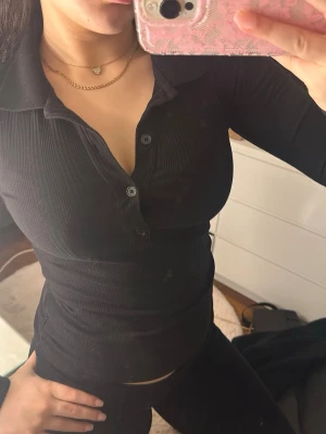 Svart ribbad topp med krage - Säljer en svart, ribbad långärmad topp med krage och knappar framtill. Figurnära passform och stretchigt material som ger en snygg siluett. Perfekt till jeans eller kjol för en trendig look.