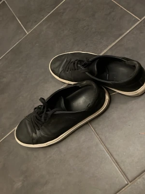 Svarta sneakers från Selected Homme - Svarta sneakers i läder från Selected Homme med klassisk låg profil och vita sulor. Skorna har svarta skosnören och en stilren design som passar till det mesta. Perfekta för dig som gillar en clean och enkel look.