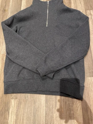 Mörkgrå halvzip hoodie - Mörkgrå hoodie med halv dragkedja framtill och hög krage. Tillverkad i mjukt sweatshirtmaterial med ribbade muddar vid ärmslut och nederkant. Enkel och stilren design utan synligt märke eller tryck, perfekt för en avslappnad look.