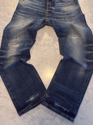 Salvage jeans från Nudie Jeans (Helt nya) - Säljer ett par blå jeans från Nudie Jeans med snygga slitningar och tvättade detaljer. Klassisk femficksmodell med orange sömmar och vågig brodyr på bakfickan. Jeansen har normal passform och är tillverkade i bomull. 