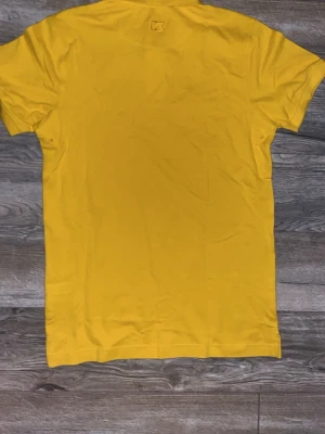 Gul pikétröja från C&A - Snygg gul pikétröja från C&A med klassisk krage och två knappar framtill. Tröjan har korta ärmar och är tillverkad i mjuk bomull, perfekt för en avslappnad och stilren look.