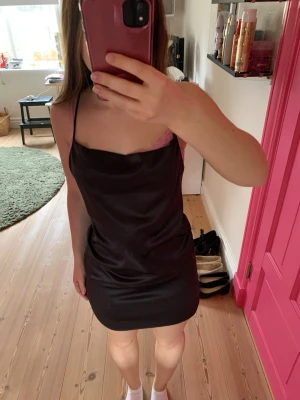 Svart kort klänning med öppen rygg - Säljer en svart kort klänning med tunna axelband och snygg öppen rygg med korsade band. Klänningen har en slät, glansig yta och en enkel, stilren design som ger en chic look. Perfekt för dig som gillar minimalistisk stil.