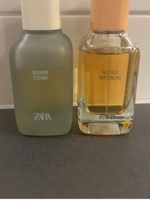 ZARA Sugar Foam & Secret Infusion parfym - Två parfymer från ZARA: Sugar Foam i en frostad, mjölkig glasflaska med vit kork och Secret Infusion i en klar glasflaska med persikofärgad kork. Båda flaskorna har en modern, stilren design och rymmer doft för dig som gillar trendiga och fräscha parfymer.