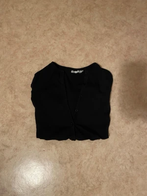 Svart basic kofta med knappar - En enkel svart kofta med rund hals och knäppning framtill. Modellen är kortare och har långa ärmar. Perfekt att slänga över en topp eller klänning för en clean look. Säljer då jag beställde i fel storlek.