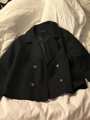 Svart skepparkavaj från Vero Moda - Stilren svart skepparkavaj från Vero Moda med dubbelknäppning och bred krage. Kortare modell med slät yta och fodrad insida. Klassisk design som passar till många olika looks. Materialet är mjukt och följsamt.