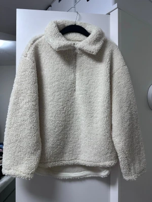 Krämvit teddyjacka med halv dragkedja - Mysig teddyjacka i krämvitt med hög krage och halv dragkedja framtill. Jackan har en oversized passform och är gjord i fluffigt, mjukt teddy-material som ger en trendig look. Perfekt för kalla dagar när du vill vara både varm och stylish.
