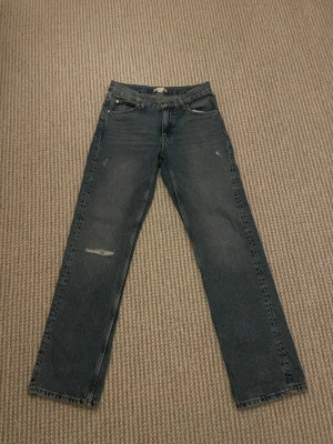 Blå raka jeans med slitningar - Säljer ett par blå raka jeans med diskreta slitningar på låret och klassisk femficksmodell. Jeansen har medel midja och är tillverkade i kraftigt denimtyg. Perfekta för en avslappnad och trendig look.