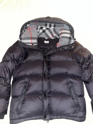 Svart pufferjacka med rutigt foder - Snygg svart pufferjacka med stor huva och rutigt foder i grått, vitt och rött. Jackan har dragkedja och knappar framtill samt två snedställda fickor med dragkedja. Perfekt för kalla vinterdagar och riktigt trendig look.