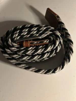 Svartvitt flätat bälte med läderdetaljer - Snyggt svartvitt bälte med tvinnat mönster och bruna läderdetaljer vid spännet och ändarna. Bältet har en metallspänne och passar perfekt för att ge din outfit en cool touch. Flexibelt material som gör det enkelt att justera.