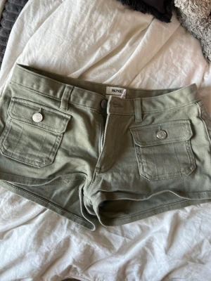 Olivgröna shorts från Monki - Snygga olivgröna shorts från Monki med två stora fickor framtill som har silverfärgade knappar. Shortsen har bälteshällor, dragkedja och knappstängning. Perfekta för varma dagar och ger en avslappnad look.