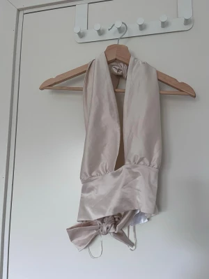 Beige omlottopp i satin - Trendig beige omlottopp i satin med breda band som knyts i midjan. Toppen är ärmlös och har en elegant, glansig finish som ger en lyxig känsla. 