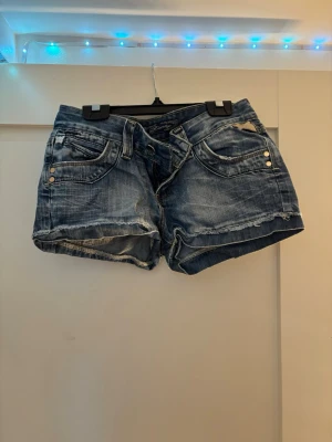 Blå jeansshorts med slitningar - Säljer ett par blå jeansshorts med slitna detaljer och råa kanter. Shortsen har fem fickor, knappgylf och dekorativa nitar fram. Köpte det secendhand