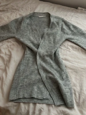 Ljusgrå ribbstickad kofta - Mysig ljusgrå ribbstickad kofta med lång ärm och öppen front. Koftan är midilång och har en smickrande passform. Perfekt att slänga över en t-shirt eller topp för extra värme. Materialet ser ut att vara mjukt och stretchigt, troligen en blandning av syntet och ull.