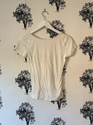 Vit topp med rosett och v-ringning - Söt vit topp med korta ärmar och djup v-ringning framtill. Toppen har en stor satinrosett som detalj vid urringningen och är tillverkad i mjuk bomull. Perfekt för en trendig och feminin look. Finns en fläck längst nere fram på toppen men går att vika upp lite!💓🌸