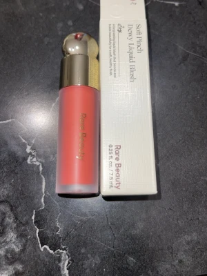 Rare Beauty Soft Pinch Dewy Liquid Blush - Flytande rouge från Rare Beauty i nyansen Soft Peach. Kommer i en frostad glasflaska med guldfärgat lock och applikator. Ger en fräsch, daggig finish och är enkel att blanda ut för ett naturligt resultat. Perfekt för dig som vill ha en mjuk persikoton på kinderna.