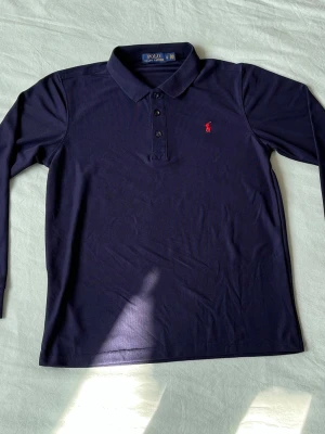 Mörkblå långärmad piké Ralph Lauren - Snygg mörkblå långärmad pikétröja från Ralph Lauren med klassisk krage och tre knappar framtill. Ikoniska röda logotypen broderad på bröstet. Tillverkad i mjukt bomullsmaterial som känns skönt mot huden.