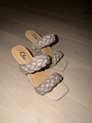 Beige pumps med flätade band från Chou - Säljer ett par snygga pumps från Chou i beige med två breda, flätade band i glittrigt material. Skorna har fyrkantig tå och låg klack, vilket ger en trendig och modern look. Perfekta för dig som vill sticka ut lite extra🔥✨