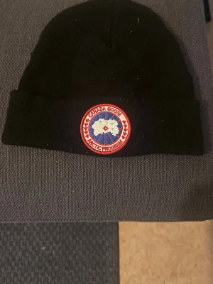 Svart Canada Goose mössa - Svart ribbstickad mössa från Canada Goose med klassisk rund logotyp-patch framtill. Perfekt för kalla dagar och ger en cool look. Materialet är troligtvis ull eller akryl och mössan har uppvikt kant.