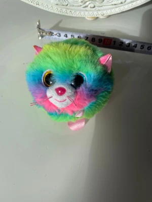 Regnbågsfärgad mjuk bollkatt - Söt bollformad mjukiskatt i regnbågsfärgad plysch med stora glittriga ögon, rosa nos och öron i satin. Katten har broderat leende och små morrhår. Perfekt som dekoration eller leksak.