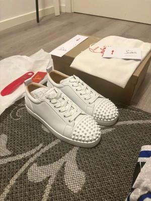 Vita sneakers från Christian Louboutin - Säljer ett par vita sneakers från Christian Louboutin i läder med coola nitar över tån. Skorna har klassisk låg profil, vita snören och en stilren design som sticker ut med sina pärlliknande detaljer framtill.