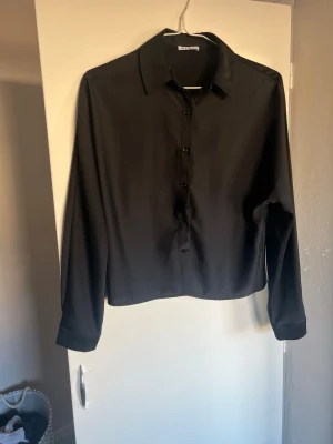 Svart klassisk skjorta med knappar - Stilren svart skjorta med klassisk krage och långa ärmar. Skjortan har knäppning framtill och är tillverkad i ett mjukt, slätt material som ger en snygg look. Perfekt för dig som gillar minimalistisk stil.