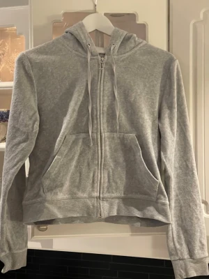 Ljusgrå zip hoodie med huva från Juicy Couture i storlek M - En stilren ljusgrå hoodie från Juicy Couture med dragkedja framtill och två stora fickor. Storlek M. Tröjan har justerbar huva med snören och ribbade muddar vid ärmar och nederkant. Perfekt för en avslappnad och bekväm look. Materialet känns mjukt och skönt mot huden.