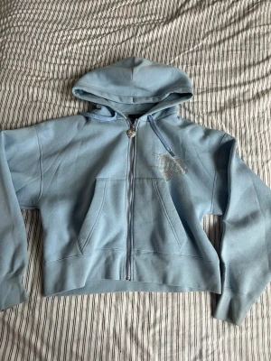 Ljusblå Juicy Couture hoodie - Säljer en ljusblå hoodie från Juicy Couture med dragkedja och huva. Framtill finns en ficka och ett glittrigt kronmotiv med texten 'Juicy' i strass. Dragsko med silverdetaljery och hjärtformad dragkedja. Perfekt för dig som gillar en trendig och bekväm stil.