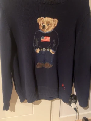 Mörkblå stickad tröja med björn från Ralph Lauren - Mörkblå stickad tröja från Ralph Lauren. Kommer passa perfekt nu till våren och man kan matcha den med nästan allt