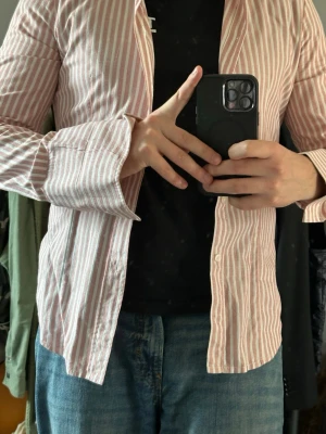 Randig skjorta i bomull - Säljer en långärmad skjorta med smala vita och ljusrosa ränder. Skjortan har klassisk krage, knäppning framtill och en bröstficka. Tillverkad i mjuk bomull, perfekt för en avslappnad men ändå stilren look.