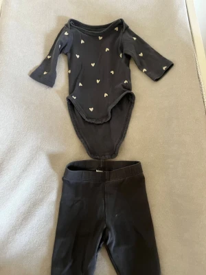 Mörkgrå body med vita hjärtan - Söt mörkgrå body med långa ärmar och vita hjärtmönster. Bodyn har praktiska tryckknappar i grenen för enkel påklädning. Tillverkad i mjukt bomullstyg som känns skönt mot huden. Perfekt för dig som gillar gulliga detaljer!