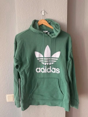 Adidas originals - En grön hoodie från adidas originals i strl 38.  Tröjan är i gott skick. Den anmärkning jag kan hitta är en diffus prick/fläck på fickan (se bild 4) som jag inte har provat behandla.