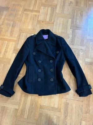 Svart skepparkavaj med dubbelknäppning - Stilren svart skepparkavaj med klassisk dubbelknäppning och bred krage. Jackan har dekorativa axelklaffar, fickor i sidorna och justerbara remmar vid ärmsluten. Perfekt för dig som vill ha en tidlös och snygg look till hösten eller vintern. Stolek 42 men är som M