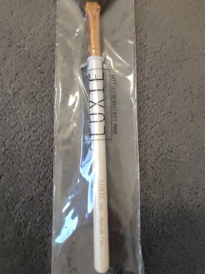 Luxie 560 Medium Fan sminkborste - En stilren sminkborste från Luxie, modell 560 Medium Fan. Borsten har ett vitt skaft med guldiga detaljer och mjuka syntetiska strån. Perfekt för att applicera highlighter eller svepa bort överflödigt puder. Kommer i originalförpackning.