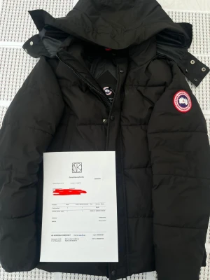 Svart dunjacka från Canada Goose - Säljer en svart dunjacka från Canada Goose med huva och klassisk logotyp-patch på ärmen. Jackan är vadderad och har dragkedja samt knappar framtill. Perfekt för kalla vinterdagar och riktigt varm tack vare dunfyllningen.