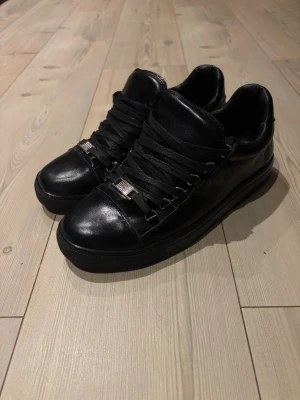 Svarta Arenas - Stilrena svarta sneakers i läder med breda svarta skosnören och metall-detaljer framtill. Klassisk låg modell med diskret design och svarta sulor. Perfekta för dig som gillar minimalistisk och clean stil.