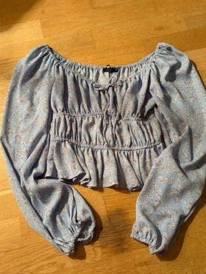 Blommig offshoulder blus från BikBok - Söt ljusblå offshoulder blus från BikBok med små blommor i pastellfärger. Blusen har långa puffärmar med resår vid ärmslut, smockdetaljer över bröstet och en dekorativ knytning framtill. Tillverkad i ett lätt och luftigt tyg.