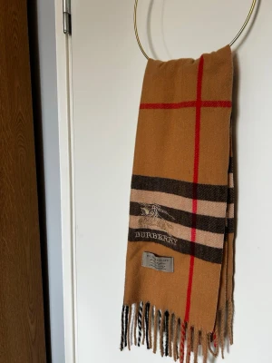 Burberry halsduk - Snygg beige halsduk från Burberry med det ikoniska rutmönstret. Halsduken har fransar i ändarna och är tillverkad i mjuk kashmir. Perfekt accessoar för att lyfta din outfit med en tidlös och lyxig känsla. Pris kan diskuteras 