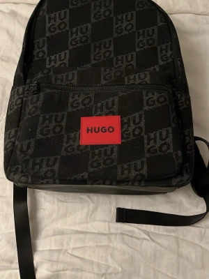 Svart ryggsäck från HUGO - Snygg svart ryggsäck från HUGO med all-over logomönster och en röd patch framtill. Väskan har en stor ytterficka med dragkedja och justerbara axelband, varav ett är rött med HUGO-tryck. Tillverkad i slitstarkt tygmaterial, perfekt för skolan eller stan.
