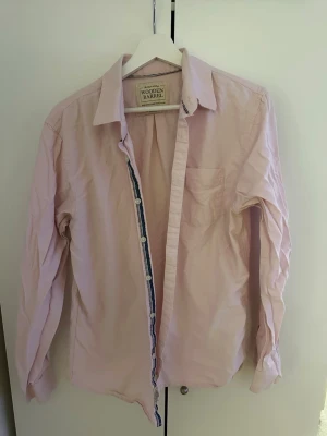 Ljusrosa skjorta från Wooden Barrel - Stilren ljusrosa skjorta från Wooden Barrel med klassisk krage, bröstficka och knäppning framtill. Skjortan har långa ärmar och är tillverkad i mjuk bomull, perfekt för en clean och avslappnad look.