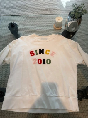 Vit sweatshirt från By Malina - Vit sweatshirt från By Malina med färgglada bokstäver som stavar 'SINCE 2010' på bröstet. Tröjan har rund halsringning och är tillverkad i mjukt bomullsmaterial. Perfekt för en avslappnad och trendig look.