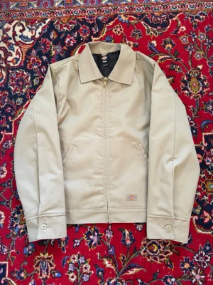 Beige Dickies Eisenhower Jacka - Klassisk beige jacka från Dickies Icons med dragkedja framtill och två snedställda fickor. Jackan har en enkel krage, knappar vid ärmsluten och quiltat svart foder på insidan. Perfekt för dig som gillar stilren och tidlös design.