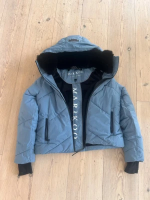 Marikoo jacka blå - Snygg, populär blå dunjacka från Marikoo med stor huva och svart fluff på insidan av kragen. Jackan har två dragkedjeförsedda fickor, quiltad design och ribbade muddar vid ärmsluten. Perfekt för kalla vinterdagar och riktigt trendig look. Endast använd några få gånger och inga skavanker!