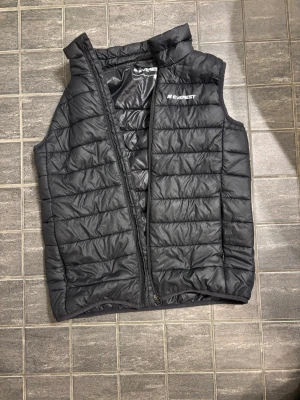 Svart dunväst från Everest - Snygg svart dunväst från Everest med quiltad design och hög krage. Västen har dragkedja framtill och diskret logga på bröstet. Perfekt för lager-på-lager och håller dig varm under kyliga dagar. Materialet är lätt och smidigt, vilket gör den enkel att bära.