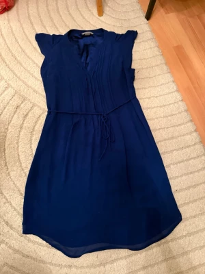 Marinblå kortklänning med knytband - Snygg marinblå kortklänning med v-ringning och tunna knytband i midjan. Klänningen har korta ärmar och lätt volang vid axlarna. Tillverkad i ett mjukt, tunt material som ger ett fint fall. Perfekt för dig som gillar enkel men stilren design.