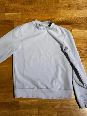 Ljusgrå sweatshirt från Lacoste S/M - Ljusgrå sweatshirt från Lacoste med diskret logga broderad på bröstet och ribbade muddar vid ärmslut och nederkant. Rund halsringning med textdetalj på insidan. Perfekt för en clean och stilren look.