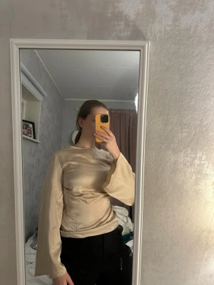 Beige satinblus med vida ärmar - Stilren beige blus i satinliknande material med rund halsringning och långa, vida ärmar. Snyggt figursydd passform som ger en elegant siluett. Perfekt för dig som gillar en trendig och minimalistisk look.