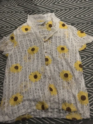 Vit blommig skjorta med spetsdetaljer - Säljer en vit skjorta med korta ärmar och stora gula blommor broderade över hela plagget. Skjortan har spetsdetaljer, knappar framtill och en avslappnad passform. Perfekt för dig som gillar unika och somriga plagg!