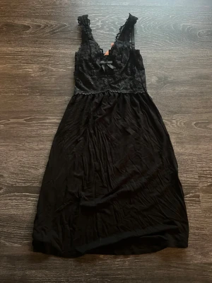 Svart spetsnattlinne från Hunkemöller - Snyggt svart nattlinne från Hunkemöller med spetsdetaljer upptill och tunna axelband. Överdelen har vacker spets och en liten rosett framtill, medan kjoldelen är i mjukt, följsamt tyg. Perfekt för en mysig kväll hemma. Aldrig använt.