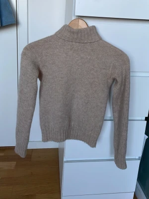 Beige polotröja i ullmix - Mysig beige stickad polotröja med ribbade muddar och hög krage. Tröjan har en klassisk rak passform och är tillverkad i mjuk ullmix som håller dig varm under kyliga dagar. Perfekt att styla med jeans eller kjol för en enkel och trendig look. Tröjan är i ett marial som är stikigt och är väldigt tajt.
