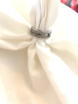 Silverfärgad ring med hjärtslag och hjärta - Cool silverfärgad ring i metall med ingraverat hjärtslag och ett hjärta. Ringen har en matt finish och är bred, vilket ger en modern och stilren look. Perfekt för dig som gillar unika detaljer och vill sticka ut lite extra.